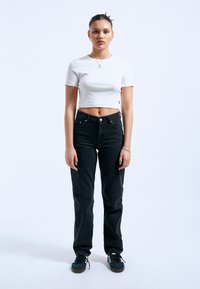 T-shirt bianco corto, jeans neri a vita alta e sneaker nere. Il modello è in posizione eretta su uno sfondo bianco semplice.