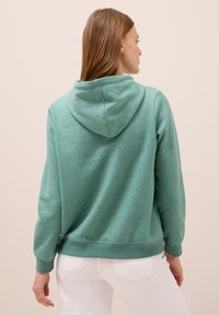 Turkos hoodie med en avslappnad passform, långa ärmar och huva på baksidan. Mjuk textur och ribbade ärmslut. Matchas med vita byxor.
