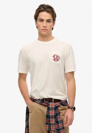 Biała bawełniana koszulka z czerwonym logo na kieszeni. Nosi z khaki spodniami i niebieską koszulą w kratę zawiązaną w talii. Zegarek na nadgarstku.