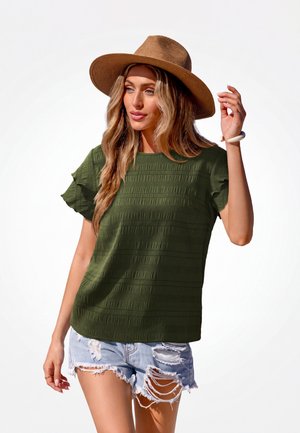 Vrouw met een bruine hoed met brede rand, olijfgroen textuur kort mouw top, gescheurde lichte denim shorts en een wit armbandje aan haar rechterpols.