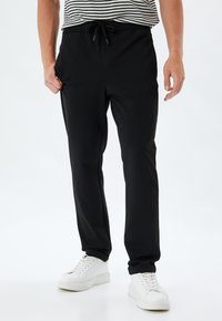 Sort joggebukser lavet af blødt stof med elastisk talje, justerbar snor og sidelommer; parret med hvide sneakers.