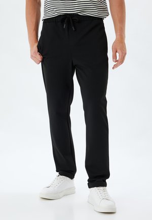 Sort joggebukser lavet af blødt stof med elastisk talje, justerbar snor og sidelommer; parret med hvide sneakers.