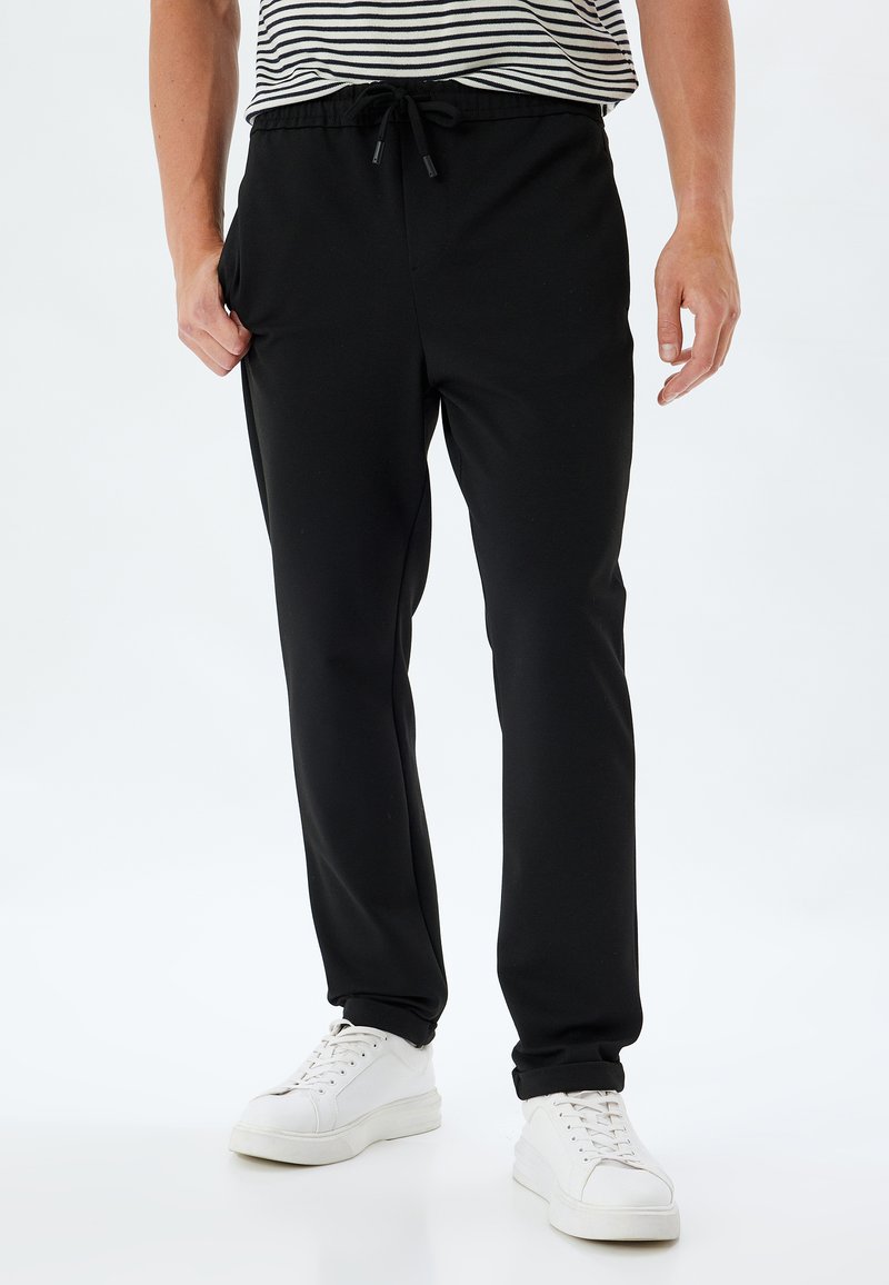 Sort joggebukser lavet af blødt stof med elastisk talje, justerbar snor og sidelommer; parret med hvide sneakers.
