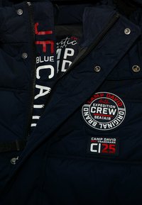 Donkerblauwe jas met ritssluitingen en drukknopen, met merkpatches van "Camp David Expedition Crew" en "CI 25" en bedrukte binnenvoering.