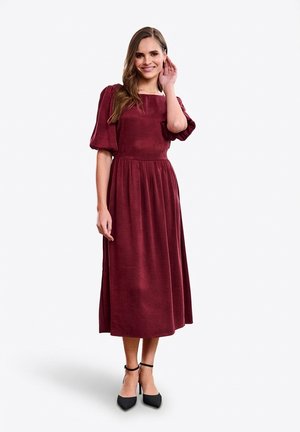 Bordowa sukienka do kolan z bufkami na rękawach, kwadratowym dekoltem i marszczoną talią. Para czarnych szpilek z paskami wokół kostek.