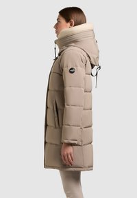 Frau in beiger, gesteppter Winterjacke mit hohem Lammfellkragen und Kapuze, stehend im Profil vor einem schlichten Hintergrund.