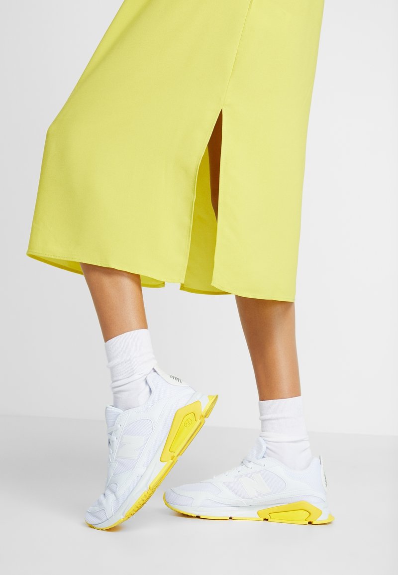 Baskets blanches avec des accents jaunes, en matériaux en mesh et synthétiques, avec des semelles plates et des lacets. Portées avec des chaussettes blanches et une robe jaune vif.