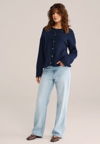 Tamsiai mėlynas megztas megztinis su tekstūruotu audiniu, raukšlėtu sagų užtrauktuku, plačiomis rankovėmis, derinamas su šviesiai mėlynomis platėjančiomis džinsų kelnėmis ir rudais smailiais bateliais.