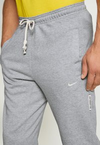Pantaloni de trening gri cu talie elastică, șnur, buzunare laterale și logo-ul alb Nike. Material moale cu o textură netedă.