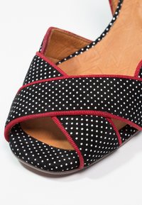 Chaussure en daim noir à pois blancs, avec des bordures rouges, un bout ouvert et des brides croisées. L'intérieur est en cuir marron clair.