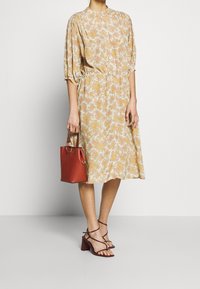 Krémové šaty s květinovým vzorem, balónovými rukávy a páskem v pase. Doplňují je červená kabelka a sandály na podpatku se řemínky.