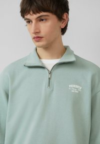 Light green half-zip sweatshirt made of soft fabric, featuring a small white logo and a textured collar with silver zipper hardware.