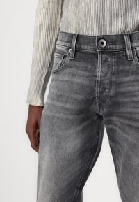 Primer plano de una persona vistiendo jeans negros desteñidos y una camiseta de manga larga gris claro con textura acanalada, con la mano izquierda apoyada a un lado.