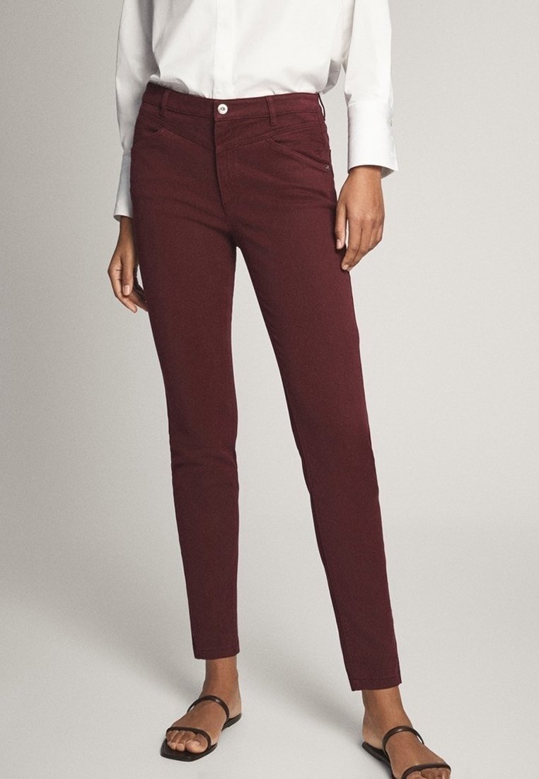 Femme portant un pantalon slim bordeaux, une chemise blanche à manches longues et des sandales noires à bouts ouverts, debout devant un fond uni.