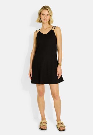 Mujer con cabello rubio corto, vistiendo un vestido negro sin mangas con doble tirante y sandalias beige, de pie contra un fondo blanco.