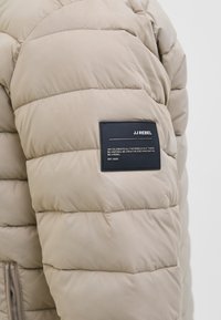 Chaqueta de plumas beige con secciones acolchadas, que presenta una etiqueta de tela negra en la manga, con el nombre de la marca y detalles en texto.