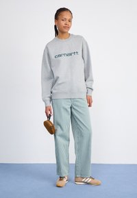 Grå sweatshirt med grøn "carhartt." tekst, parret med lyseblå og hvide stribede brede bukser og en brun taske. Beige sneakers.