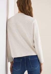 Femme portant une chemise beige à manches longues et un jean bleu, de dos, ses cheveux visibles, sur un fond clair uni.