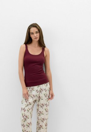 Frau mit langen braunen Haaren, die ein ärmelloses burgunderfarbenes Top und weiße Hosen mit Blumenmuster trägt, steht vor einer einfachen weißen Wand.