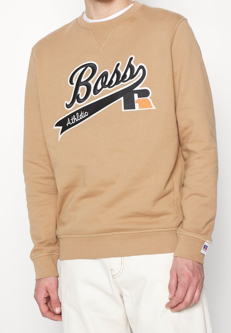 Sweat beige avec logo "Boss Athletic" noir et blanc, porté avec un pantalon blanc, montrant le haut du torse et la main droite.