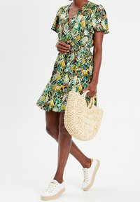 Robe à imprimé floral en vert, jaune et marron. Décolleté en V, manches courtes, taille cintrée. Sac à main en osier naturel et baskets blanches avec semelles beiges.