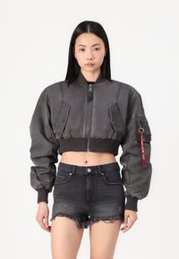 Gray cropped bomber jacket with a front zipper, ribbed cuffs, shoulder patches, and decorative tag. Worn with black distressed denim shorts.