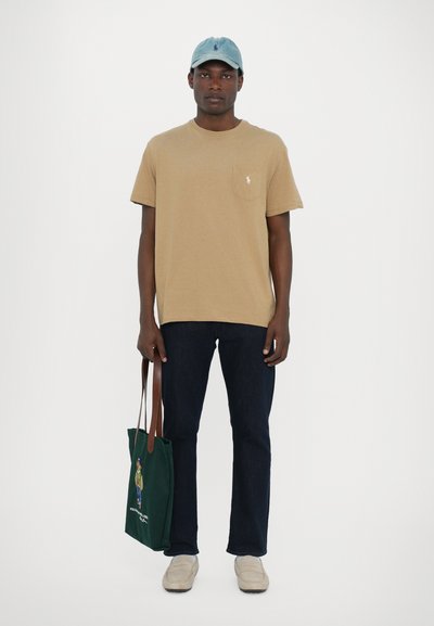 T-shirt beige à manches courtes avec une poche poitrine, jeans bleu foncé, chaussures slip-on beige clair et un sac fourre-tout vert avec un design coloré.