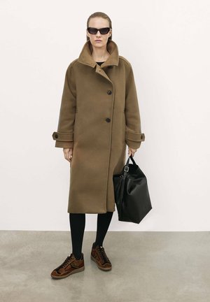 Cappotto di lana marrone con ampio colletto e chiusura a bottoni, abbinato a collant neri e sneakers marroni a fantasia. Tiene una borsa tote nera.