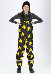 Schwarze wasserdichte Latzhose mit gelben Gummiente-Mustern, verstellbaren Trägern und reflektierenden Akzenten an den Beinen. Schwarze Stiefel getragen.