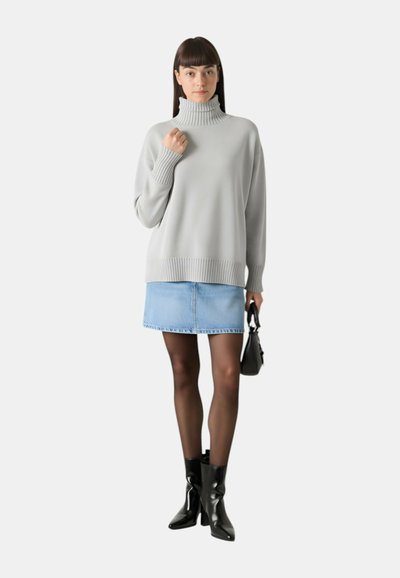 Maglione color grigio chiaro a collo alto con polsini e orlo a coste, abbinato a una gonna di denim blu e stivali neri alla caviglia. Borsa nera semplice.