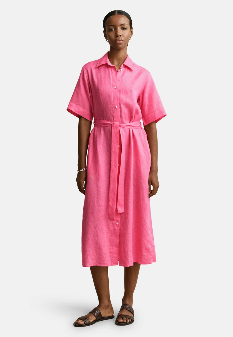 Femme portant une robe midi boutonnée rose vif avec une ceinture à la taille, des manches courtes et des sandales plates marron, debout devant un fond uni.