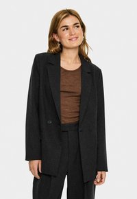 Donna con un blazer doppiopetto grigio scuro e pantaloni abbinati, indossando un top marrone aderente, sorride leggermente guardando in alto e a destra.