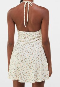 Kvinna klädd i en ärmlös vit klänning med knytning bak och små rosa och gröna blommönster, sedd bakifrån mot en enfärgad bakgrund.