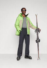 Chaqueta de esquí de color verde brillante con detalles en beige, superpuesta a una camisa gris, combinada con pantalones de esquí negros y botas de esquí negras, sosteniendo esquís.
