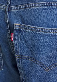 Blå denimjeans med en distinkt bakficka som har gul sömnad och en röd Levi's-tag. Tyget har en klassisk texturerad finish.