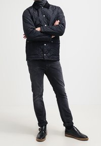 Kvadra svart jacka med krage och tryckknappar, i kombination med mörka jeans och svarta skor. Enkel design utan synliga mönster.