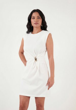 Kvinde med mørkt hår iført en ærmeløs hvid kjole med en guldfarvet spænde detalje i taljen, stående mod en ensfarvet baggrund.