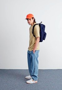 Brun t-shirt, blå baggy jeans, vita sneakers med orange detaljer, och en klar orange keps. En marinblå ryggsäck hänger över ena axeln.