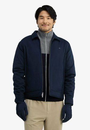 Homme portant une veste bleu marine, un pull gris à fermeture éclair, un pantalon beige et des gants en tricot bleu marine, debout et souriant sur fond blanc.