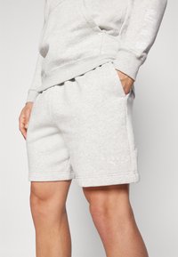 Pantalones cortos de sudadera grises, hechos de un tejido suave, con cintura elástica, bolsillos laterales y un logo blanco en la pierna izquierda. Diseño simple.