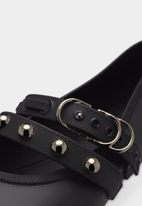 Chaussure plate noire avec deux brides ; une bride est ornée de clous argentés, l'autre comporte deux boucles argentées.