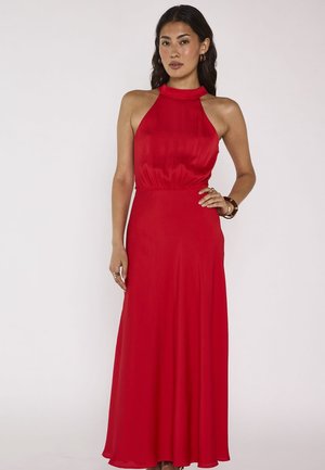 Femme debout devant un fond uni, portant une robe rouge longue sans manches à col halter, avec des accessoires minimalistes.