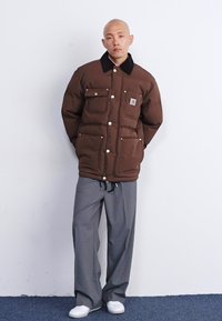 Chaqueta acolchada marrón con un collar negro, múltiples bolsillos frontales, asegurados con botones a presión, combinado con pantalones de pierna ancha grises y zapatos blancos.