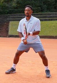 Tenisový hráč stojí na antukovom kurte, oblečený v svetlomodrej polokošeli a šortkách, s bielymi zápästnými páskami a sivými tenisovými topánkami, držiac raketu.