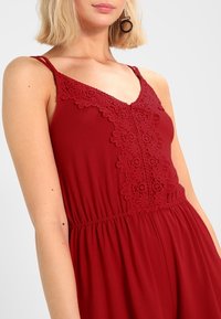 Röd ärmlös jumpsuit med spetsdetaljer längs halsringningen, elastisk midja och mjukt tyg. Har tunna axelband.