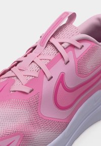 Prim-plan cu o pantof sport roz, cu material din plasă, șireturi, o buclă pentru tragere și un swoosh Nike într-o nuanță mai închisă de roz pe lateral.