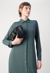 Robe à manches longues et boutons en tissu vert foncé. Le modèle tient un sac à main plissé noir texturé. Design de col simple, texture douce.