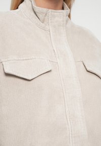 Veste en velours côtelé beige clair avec col montant, deux poches poitrine et texture côtelée verticale. Comprend une fermeture éclair centrale.