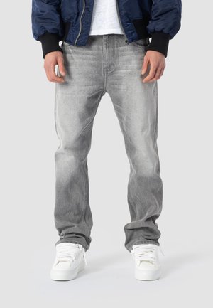 Mann trägt ausgewaschene graue Jeans, weiße Turnschuhe, ein weißes Hemd und eine marineblaue Jacke mit schwarzen Bündchen und steht vor einem schlichten Hintergrund.