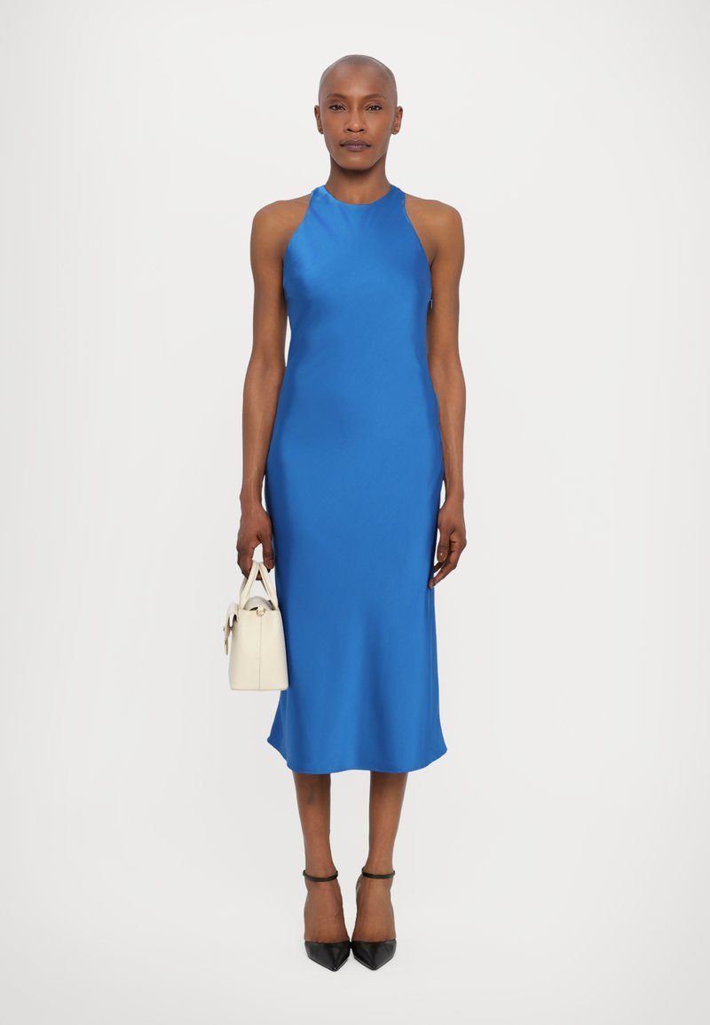 Robe nuisette en satin bleu avec un col haut et sans manches. Portée avec des escarpins noirs à bout pointu et un petit sac à main blanc.
